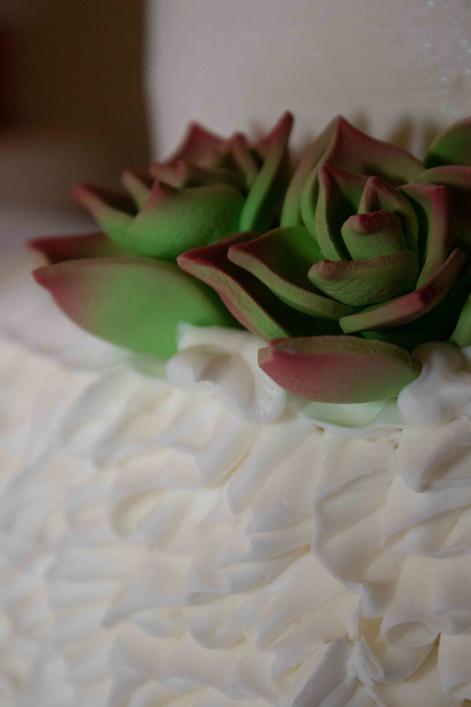 Close-up of a wedding cake. Frosting is visible on the bottom, while a green flower with red tips dominates the top of the cake.
