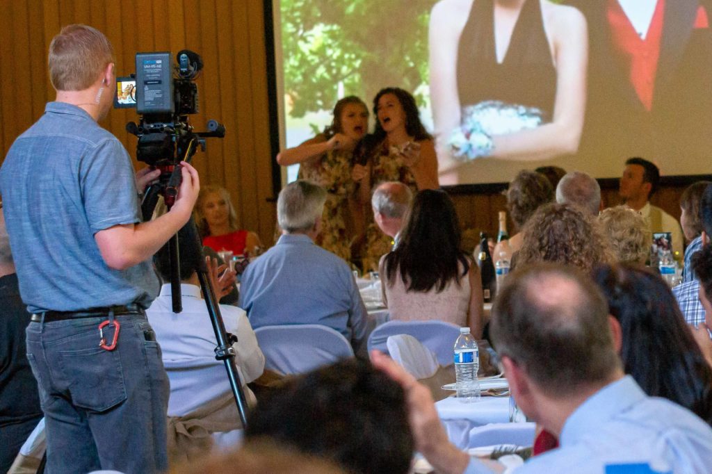 Image of a wedding party. A cameraman stands in the middle of crowded tables while two women are speaking.
